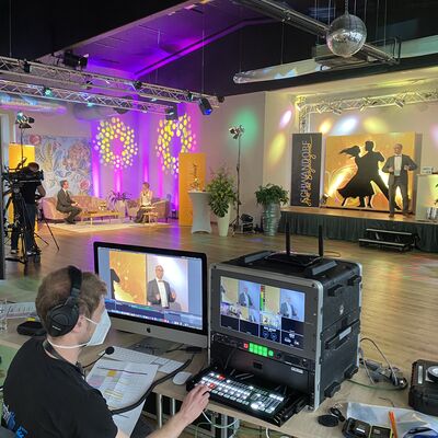 Foto von der Technik, die hinter dem Live-Stream steckt.
2 Bildschirme, viele Tasten und eine Person. Im Hintergrund sieht man die dekorierten Rume der Tanzschule Theuerl.