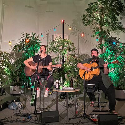 Die Musiker Susi Raith und Mathias Kellner spielen auf eine Bhne Gitarre und singen. Die Bhne ist mit vielen Pflanzen und bunten Lichtern dekoriert.