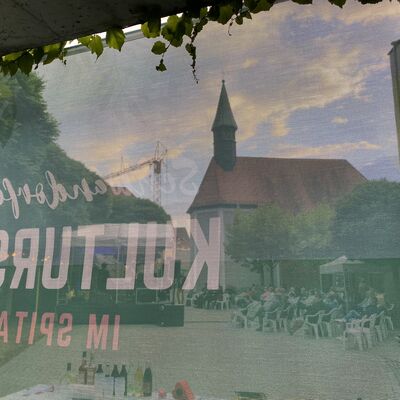 Spitalkirche, Bhne im Spitalgarten und das Publikum davor durch ein Werbetransparent hindurch bei Dmmerung fotografiert.