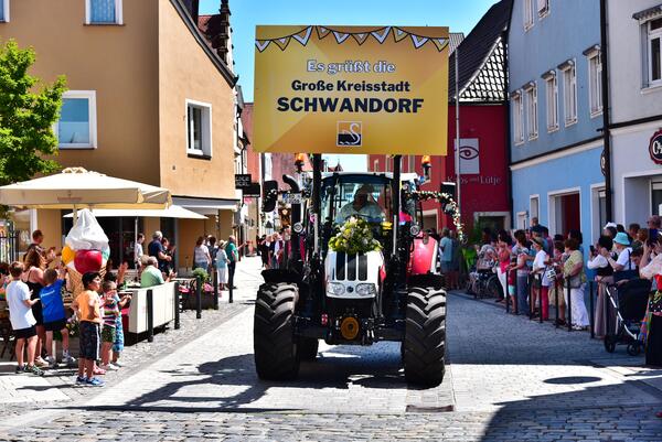 Bild vergrößern: Ein Bulldog fhrt als Teil eines Festzuges die Strae entlang. Darauf ist ein groes gelbes Schild mit der Aufschrift "Es grt die groe Kreisstadt Schwandorf" montiert.