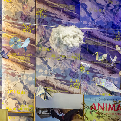 Vor einer mit Zwickl-Plakaten beklebten Wand hngen an einer Schnur eine Runde Wolke aus Watte, Origami-Vgel und CDs.