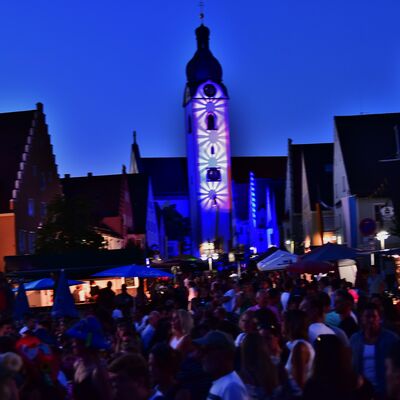 Bild vergrößern: Der mit einem blau-weiem Muster illuminierte Kirchturm St. Jakob beim Schwandorfer Brgerfest. Davor sind umrisshaft viele Menschen auf dem Marktplatz zu sehen.