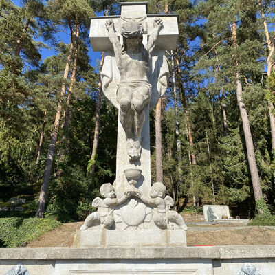 Ein groes Kreuz aus Stein mit zwei Engeln, welche Jesus zu Fen liegen und einen Kelch zu ihm emporheben. Im Hintergrund stehen viele Bume.