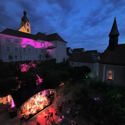 Bild vergrößern: Luftaufnahme der Volkshochschule Schwandorf, die pink angeleuchtet wird. Unterhalb, im Hof des Spitalgartens steht eine Open-Air-Bhne die hellrot erleuchtet ist.