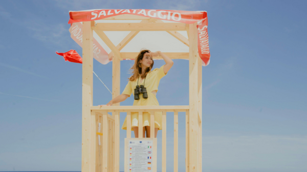 Die Sngerin Luca Vasta steht auf einem Rettungsturm am Strand und blickt in die Ferne. Um den Hals trgt sie ein schwarzes Fernglas. 