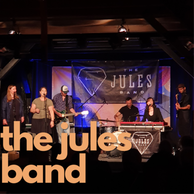 Orangene Schrift auf Foto: "the jules band". Im Hintergrund ist die Musikgruppe auf der Bhne im Sperlstadel des Come Together Festivales 2021.