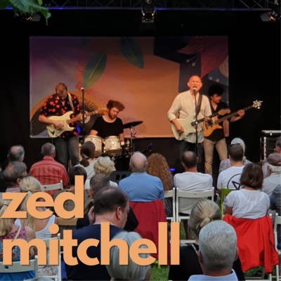 Orangene Schrift auf Foto: "Zed Mitchell"
Im Hintergrund spielt die Musikgruppe um Zed Mitchell auf der Open-Air Bhne des Come Together Festivales 2021.