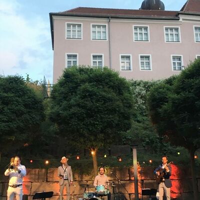 Foto: Auf einer erleuchteten Open-Air Bhne spielen 4 Musiker der Musikgruppe Bavaschoro beim Kultursommer im Spitalgarten 2020. Sommerabendliche Stimmung.