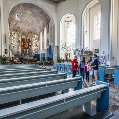 Bild vergrößern: Eine Familie mit einem Fhrer bei der Kirchenfhrung in der Kreuzbergkirche. Die Bnke in der Kirche sind hellblau und im Hintergrund sind die groen Fenster und der prchtige Altar zu sehen.